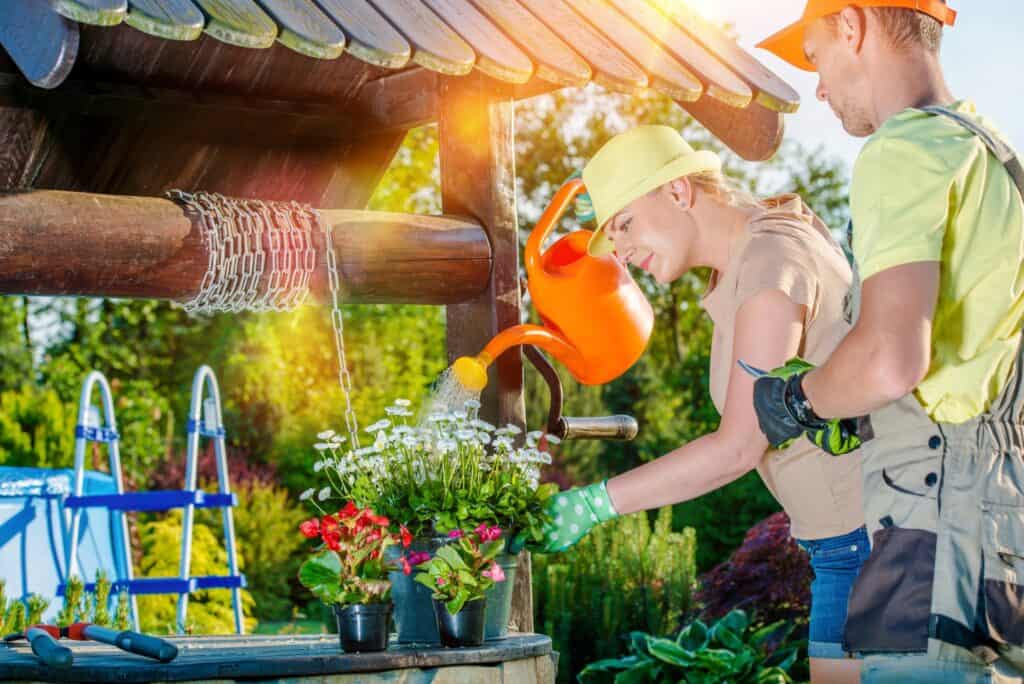 Dwie osoby w czapkach i rękawiczkach podlewają kwiaty w doniczkach przy drewnianej studni w nasłonecznionym ogrodzie. Jedna z nich trzyma konewkę, a druga pomaga w układaniu roślin.
