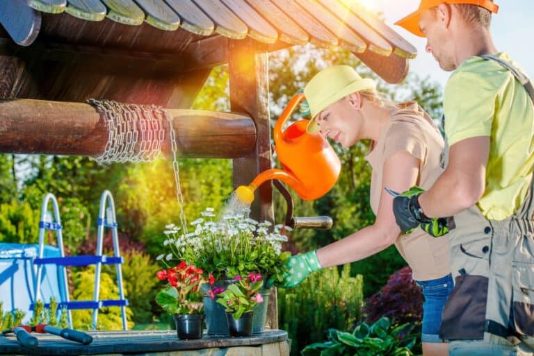 Dwie osoby w czapkach i rękawiczkach podlewają kwiaty w doniczkach przy drewnianej studni w nasłonecznionym ogrodzie. Jedna z nich trzyma konewkę, a druga pomaga w układaniu roślin.